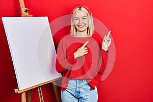 Beautiful blonde woman standing by painter easel stand smiling and looking at the camera pointing with two hands and fingers to