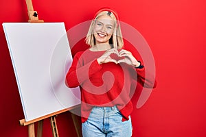 Beautiful blonde woman standing by painter easel stand smiling in love doing heart symbol shape with hands