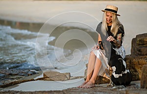 Beautiful blonde woman in straw hat sitting on a rock and petting the good dog. The same color. Thai beach. Morning walk