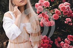 Beautiful blonde woman wear stylish white top with corset posing over blooming flower roses outdoors close up.
