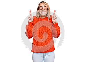 Beautiful blonde woman wearing casual clothes and glasses gesturing finger crossed smiling with hope and eyes closed