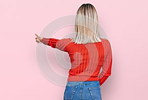 Beautiful blonde woman wearing casual clothes posing backwards pointing ahead with finger hand