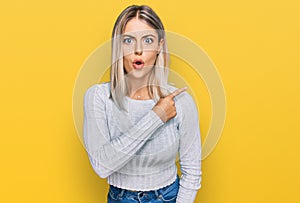 Beautiful blonde woman wearing casual clothes surprised pointing with finger to the side, open mouth amazed expression