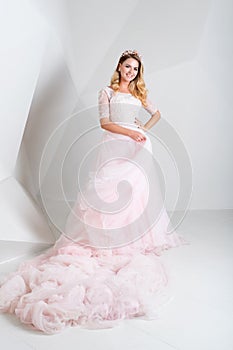 Beautiful blonde woman wearing gorgeous long dress and crystal crown posing in studio on white polygonal background.