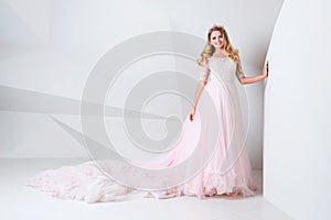 Beautiful blonde woman wearing gorgeous long dress and crystal crown posing in studio on white polygonal background.