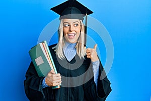 Beautiful blonde woman wearing graduation cap and ceremony robe holding books pointing thumb up to the side smiling happy with