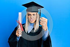 Beautiful blonde woman wearing graduation cap and ceremony robe holding degree annoyed and frustrated shouting with anger, yelling