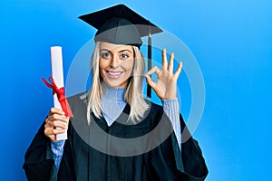 Beautiful blonde woman wearing graduation cap and ceremony robe holding degree doing ok sign with fingers, smiling friendly