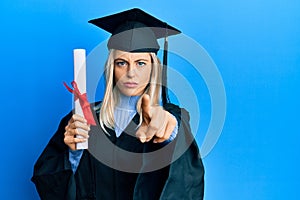 Beautiful blonde woman wearing graduation cap and ceremony robe holding degree pointing with finger to the camera and to you,