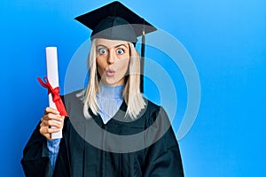 Beautiful blonde woman wearing graduation cap and ceremony robe holding degree scared and amazed with open mouth for surprise,