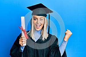 Beautiful blonde woman wearing graduation cap and ceremony robe holding degree screaming proud, celebrating victory and success