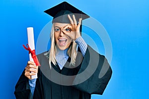 Beautiful blonde woman wearing graduation cap and ceremony robe holding degree smiling happy doing ok sign with hand on eye