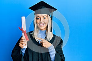 Beautiful blonde woman wearing graduation cap and ceremony robe holding degree smiling happy pointing with hand and finger