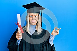 Beautiful blonde woman wearing graduation cap and ceremony robe holding degree smiling happy pointing with hand and finger to the