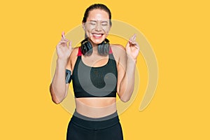 Beautiful blonde woman wearing gym clothes and using headphones gesturing finger crossed smiling with hope and eyes closed