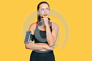 Beautiful blonde woman wearing gym clothes and using headphones with hand on chin thinking about question, pensive expression