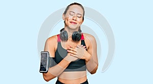 Beautiful blonde woman wearing gym clothes and using headphones smiling with hands on chest with closed eyes and grateful gesture