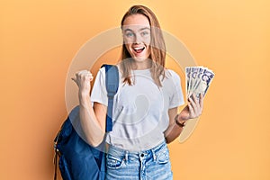 Beautiful blonde woman wearing student backpack and holding 5000 japan yen pointing thumb up to the side smiling happy with open