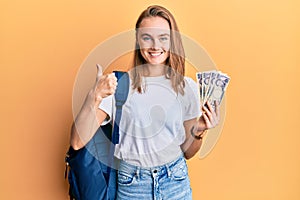 Beautiful blonde woman wearing student backpack and holding 5000 japan yen smiling happy and positive, thumb up doing excellent