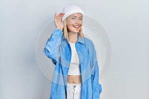 Beautiful blonde woman wearing wool hat smiling with hand over ear listening an hearing to rumor or gossip