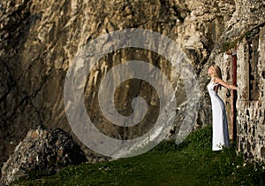 Beautiful blonde woman in white long dress outdoor, enjoys the sunlight next to the door of a ruined building in Ireland