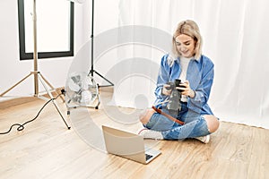 Beautiful blonde woman working as professional photographer at photography studio sitting on the floor checking photos on computer