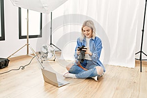 Beautiful blonde woman working as professional photographer at photography studio sitting on the floor checking photos on computer