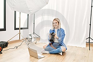 Beautiful blonde woman working as professional photographer at photography studio sitting on the floor checking photos on computer