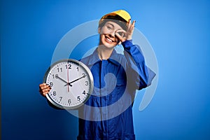 Beautiful blonde worker woman with blue eyes wearing security helmet holding big clock with happy face smiling doing ok sign with