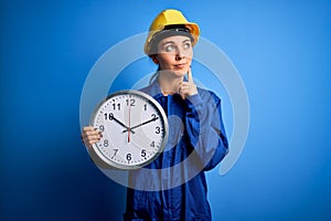Beautiful blonde worker woman with blue eyes wearing security helmet holding big clock serious face thinking about question, very