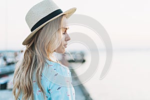 Beautiful blonde young woman in blue dress and straw hat on the pier on sunset