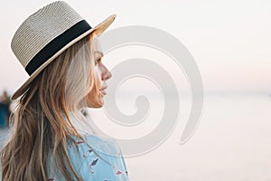 Beautiful blonde young woman in blue dress and straw hat on pier on sunset