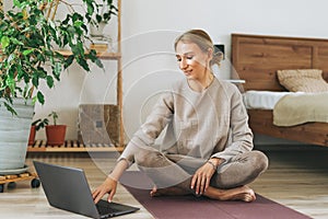 Beautiful blonde young woman in comfortable sport wear practice yoga online in cozy bedroom at home