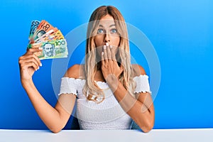 Beautiful blonde young woman holding australian dollars covering mouth with hand, shocked and afraid for mistake