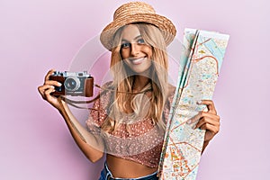 Beautiful blonde young woman holding city map and vintage camera smiling with a happy and cool smile on face