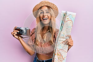 Beautiful blonde young woman holding city map and vintage camera smiling and laughing hard out loud because funny crazy joke