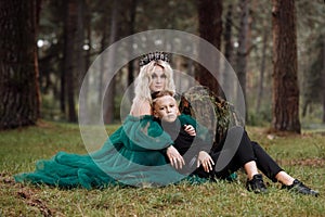 A beautiful blonde young woman in a long green dress and a diadem on her head with stylish young son in the forest. girl and boy