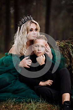 A beautiful blonde young woman in a long green dress and a diadem on her head with stylish young son in the forest. girl and boy