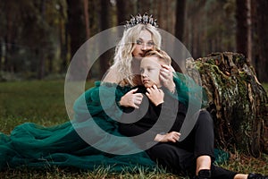 A beautiful blonde young woman in a long green dress and a diadem on her head with stylish young son in the forest. girl and boy