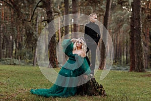 A beautiful blonde young woman in a long green dress and a diadem on her head with stylish young son in the forest. girl and boy