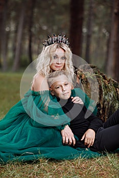 A beautiful blonde young woman in a long green dress and a diadem on her head with stylish young son in the forest. girl and boy