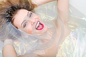 Beautiful blonde young woman with red lips laughing, screaming & relaxing laying in a bath water happy smiling