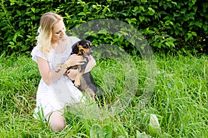 A beautiful blonde young woman sitting in green grass holds her cute dog in her arms