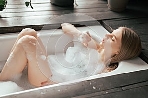 Beautiful blonde young woman taking bath with bubbles in bathroom interior with plenty green plans