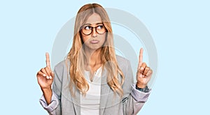 Beautiful blonde young woman wearing business clothes pointing up looking sad and upset, indicating direction with fingers,