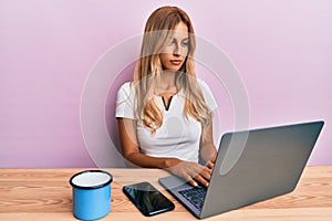 Beautiful blonde young woman working using computer laptop thinking attitude and sober expression looking self confident
