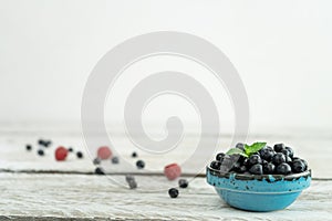 A beautiful blue bowl full of ripe blueberries stands on a light wooden table against a white wall.