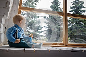 Beautiful blue-eyed baby sitting on the windowsill and looks out the window at the green trees