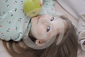 A beautiful blue-eyed girl lies with a green apple on the bed. Smiling girl.