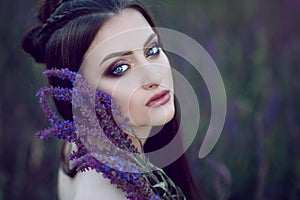 Beautiful blue-eyed lady with perfect make up and plaited hairstyle sitting in the field and holding purple flowers at her face